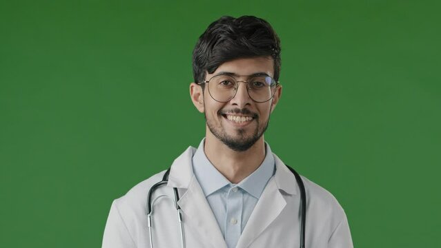 Portrait of smiling arab guy pharmacist therapist professional physician doctor wears white coat with stethoscope look at camera male medical adviser stand posing isolated in green studio background