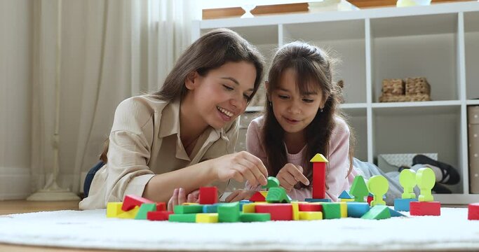 Young mother play wooden bricks with little daughter, lying on warm floor in cozy playroom looking happy enjoy playtime and leisure together at home. Child games, modern toys store ad, hobby concept