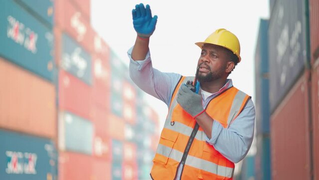 African American cargo container worker as foreman or technician use walkie talkie to comunicate with his co-worker in workplace with layer or stack of tank as background.