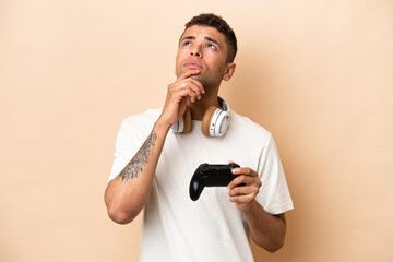 Fototapeta na wymiar Young Brazilian man playing with a video game controller isolated on beige background and looking up