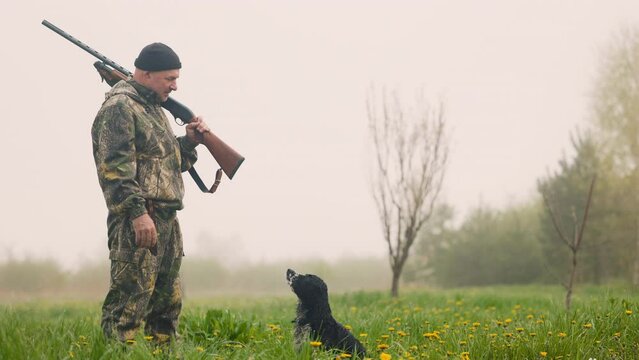 In the autumn morning, a hunter armed with a rifle and a knife stands in the background of the field. He puts the gun to cry, a hunting dog runs to him, licks. The hunter strokes the dog's head.
