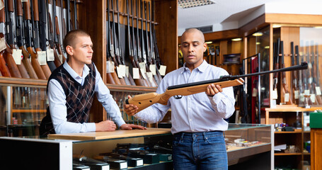 Hispanic man examining hunting break-barrel air rifle in gun shop, consulting with salesman