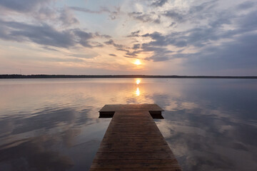 Forest lake at sunset. Wooden pier. Soft sunlight, glowing clouds, symmetry reflections in a...