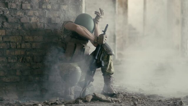 A young military Ukrainian soldier sits on the territory of an abandoned building during the fighting. An explosion occurs and dust falls from the destroyed walls. The concept of the war in Ukraine.