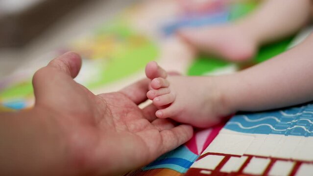 Daddy&rsquo;s hand holds little baby foot. Father caresses kid&rsquo;s tiny fingers and child moves his fingers slowly. Close up. Blurred backdrop.
