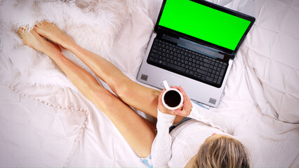 Top-down view of a woman sitting on the bed using laptop computer drinking coffee
