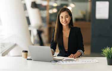 Fototapeta premium Cheerful business lady working on laptop in office, Asian happy beautiful businesswoman in formal suit work in workplace. Attractive female employee office worker smile.