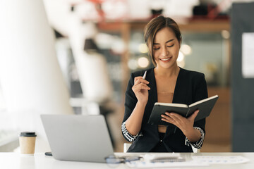 Fototapeta premium Smiling young asian businesswoman taking notes while sitting with laptop computer, Happy Smiling Girl Working Online Or Studying And Learning While Using Notebook. Freelance Work.