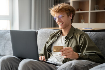 Successful freelancer ginger man holding credit card, using laptop