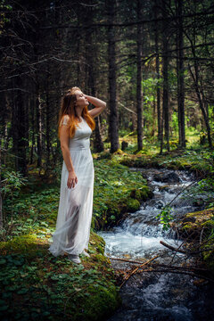 Young beautiful woman in white dress near icy stream on evergreen trees background in natural sunlight. Vertical image romantic nymph in summer forest.