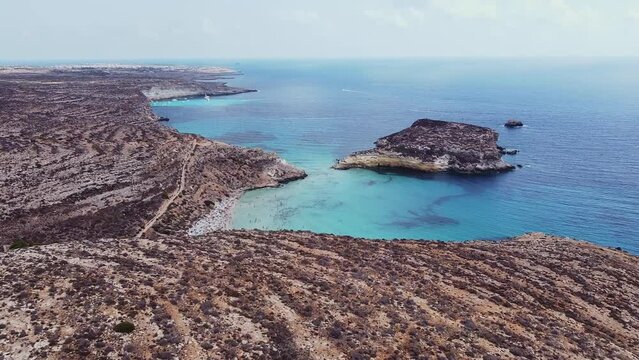 Aerial drone. Spiaggia and Isola dei Conigli, Lampedusa. Tranquil, cove-style beach with white sand and turquoise surf bordered by rocky cliffs.