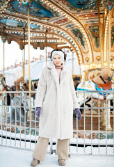 Obraz na płótnie Canvas A beautiful young woman in a faux fur coat and snake clothes poses next to a carousel at a Christmas market.
