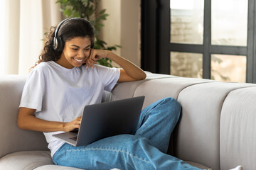 Young African American hipster woman in earphone with Afro hair using a laptop talking to webcam with friend online lying on the couch at home. Video call, virtual chat concept