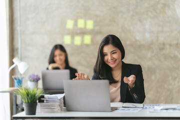 Attractive asian young confident businesswoman sitting at the office table with group of colleagues in the background, working on laptop computer.