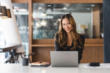 Fototapeta premium Beautiful Brunette Asian businesswoman working with a laptop at the office. Looking at camera.