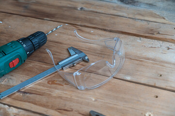 Electric drill, caliper, pliers and safety goggles lie on a wooden surface. The drill is mounted in a chuck. Selective focus.