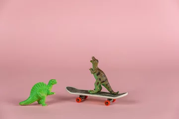 Tuinposter Two miniature dinosaurs and a mini skateboard against the pink background. Small green miniatures of predatory dinosaurs. Close-up.  Selective focus. © Mikhail