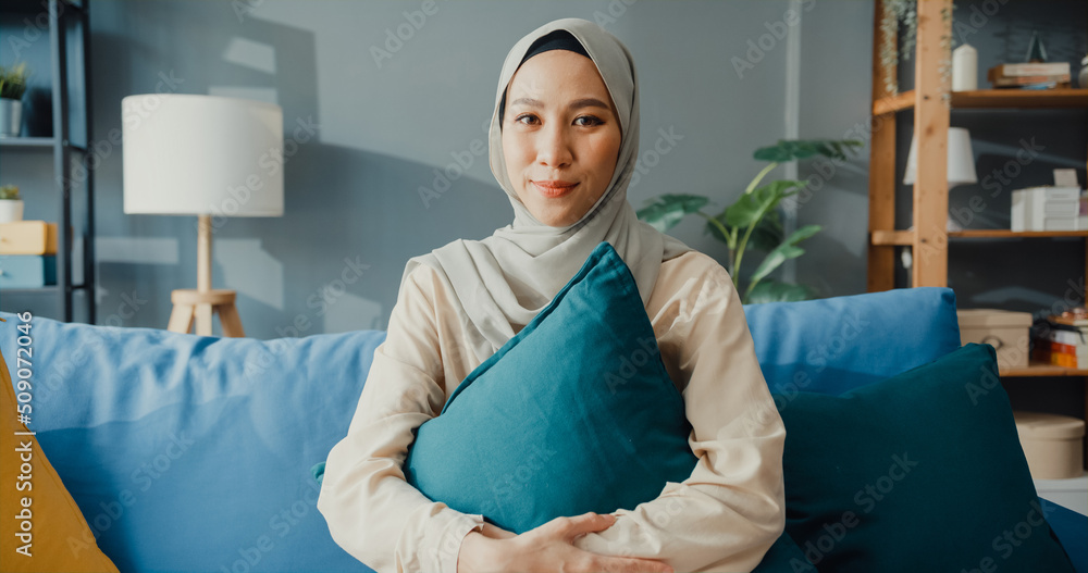 Wall mural attractive cheerful young asian muslim beauty woman in hijab with casual sitting on couch smile look