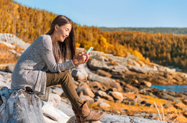 Mobile phone travel lifestyle Asian woman using cellphone data durig outdoor hiking adventure in Quebec, Canada. Happy girl texting in forest autumn foliage. Technology