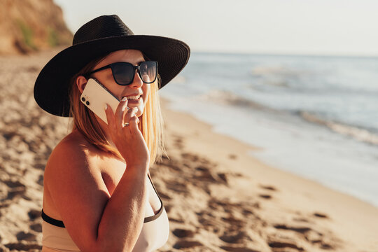 Close Up of Smiling Young Woman in Sunglasses and Hat Looking Towards the Sea While Talking on Phone
