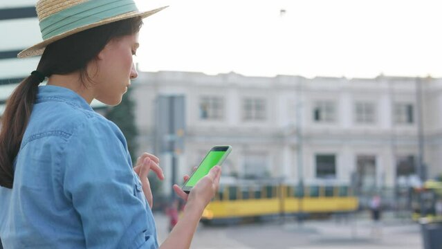 Female on bus station outdoor tapping on cellphone green screen. Woman view photos and videos on the phone. Girl using smartphone while yellow train riding at background. Technology concept.