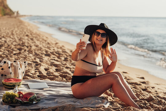Smiling Young Woman Taking Video Call on the Phone While Sitting on Sandy Beach by the Sea