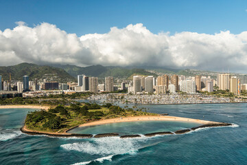 Aerial view of city tall buildings in Honolulu and Magic Island Lagoon, Hawaii. Blue sky with clouds rolling above the mountains