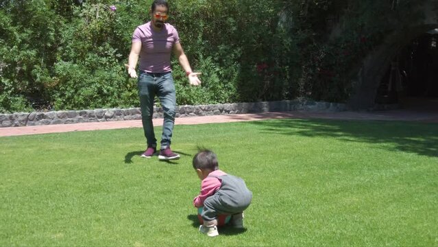 Latin dad and his baby boy playing outdoors with a ball on the grass on a sunny day