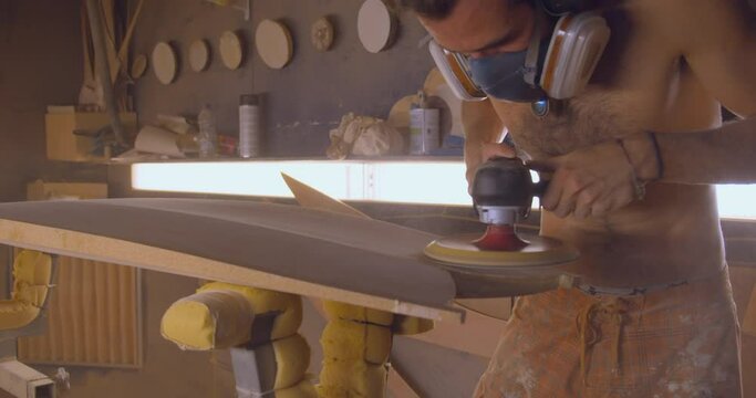 Shaper with respirator mask shaping surfboard with sanding machine. Gimbal