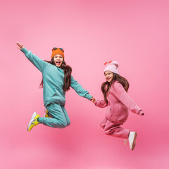 Two smiling Latin child girls in warm fashionable tracksuits jumping on a pink studio copy space. Children's beauty models with dark long hair advertising sports outerwear. Promotion of clothing store