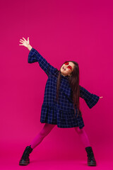 teenage girl Asian appearance, with long black hair, dressed in fashionable dress and shoes stands on bright pink background, arms wide apart. child happily smiles with an open smile and looks away.