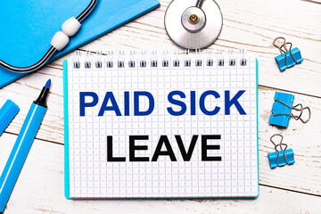 On a light wooden background, a stethoscope, a blue notepad, blue paper clips, a blue marker and a sheet of paper with the text PAID SICK LEAVE. Medical concept