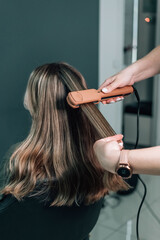 Fototapeta premium Woman hairdresser straightens brown hair of a beautiful woman using hair tongs in a beauty salon. Female stylist makes a hairstyle for a young beautiful woman in a barbershop.