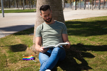 Handsome young homosexual man is sitting on the grass in the park reading a book, next to him is a gay pride flag. Concept reading, education, teaching, free time and leisure.