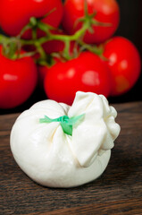 whole Italian burrata cheese lies on a wooden board, a bunch of red tomatoes can be seen in the background. Vertical snapshot.