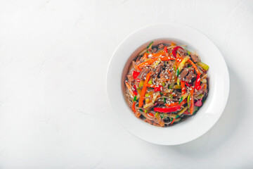 starch noodles with vegetables and beef in a white round plate on a gray background, copy space, top view.