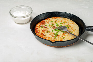 Swiss traditional dish is a potato pancake in a frying pan with a fork, decorated with green onions. there is a gravy boat with white cream sauce nearby. side view.