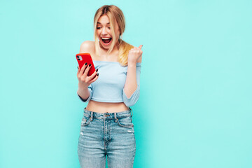 Young beautiful smiling female in trendy summer clothes. Excited and amazed woman posing near blue wall in studio. Shocked and lucky model reading modern device, celebrating holding phone