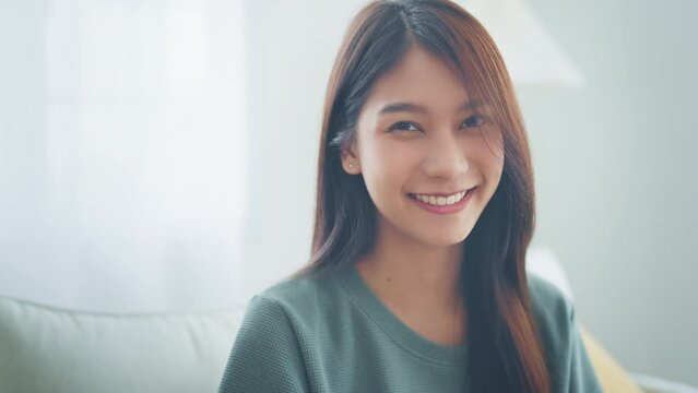 Confident smiling young adult asian woman looking at camera while sitting at home, Happy beautiful lady pretty face dental smile posing alone indoors, Slow motion close up view portrait