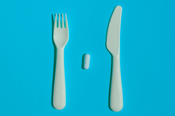A fork and a knife between which there is one white vitamin tablet on a blue background. Top view