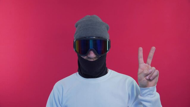 Snowboarder Shows Two Figers, Peace Sign. Man in Snowboard Goggles in a White Jacket on a Red Background in the Studio