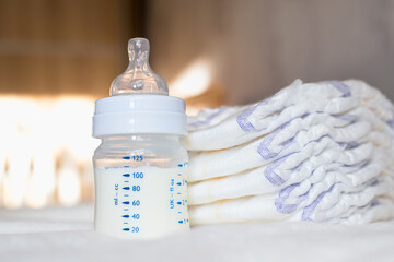 Baby bottle with milk and diapers on a white fabric background. Baby care. First days of life. Feeding and motherhood - obrazy, fototapety, plakaty