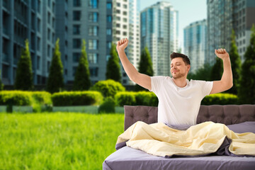 Happy man stretching in bed and beautiful view of green grass and cityscape on background. Good...