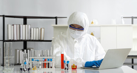 Epidemiological researcher in virus protective cloth looking at red liquid in measuring cylinder...