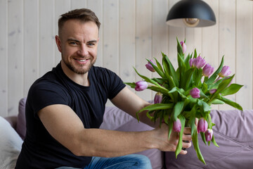 Young handsome man is sitting at home on sofa with bouquet of tulips and preparing surprise for his girlfriend. Preparing for date. Attention and care.