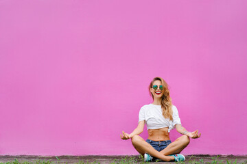 Colorful yoga. Attractive young woman sitting on lotus position on the ground against pink wall.