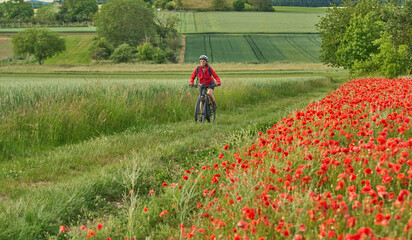 nice, active senior woman having fun on her electric bicycle in a huge field of blooming red poppies 
