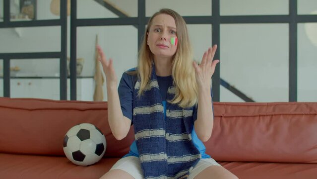 Emotional attractive middle aged woman football supporter with Italy flag painted on cheek in sport jersey watching soccer match on tv, supporting national team and nervously biting fist indoors.