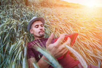 Pensive middle aged agronomist wearing hat lying on wheat field with portable tablet in hands....