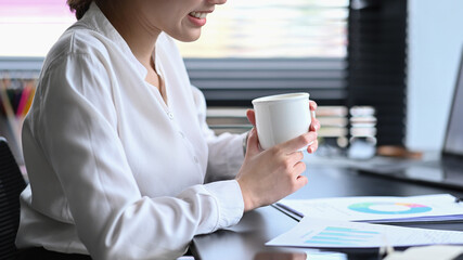 Cropped shot smiling asian female office worker sitting at her workplace and drinking hot coffee